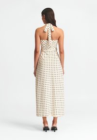 Vestido maxi de lunares en crema con puntos negros, que presenta un escote halter con lazo, un cuerpo ajustado y una falda fluida. Textura de tela suave.