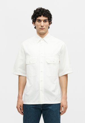 Νέος άνδρας με σγουρά σκουρόχρωμα μαλλιά φοράει λευκό κοντομάνικο πουκάμισο με δύο μπροστινές τσέπες, σε συνδυασμό με μπλε τζιν, στέκεται μπροστά σε λευκό φόντο.