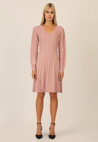 Hellrosa gestricktes Kleid mit V-Ausschnitt, langen Ärmeln, Zopfstrickmuster, weit auslaufendem Rock und gerippten Bündchen. Tragen Sie dazu schwarze Riemchensandalen.