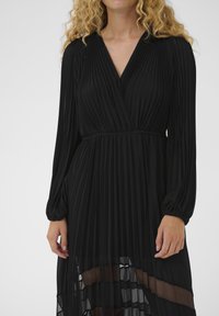 Vestido negro plisado con mangas largas, escote en V profundo, cintura elástica y paneles transparentes en el dobladillo. Tela ligera con textura suave.