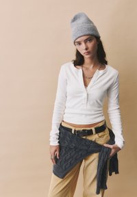 Valkoinen pitkähihainen henley-paita, jossa on osittain napitettu kaula-aukko, yhdistettynä keltaisiin samettihousuihin ja harmaaseen neulotuun huiviin, joka on sidottu vyötärölle.