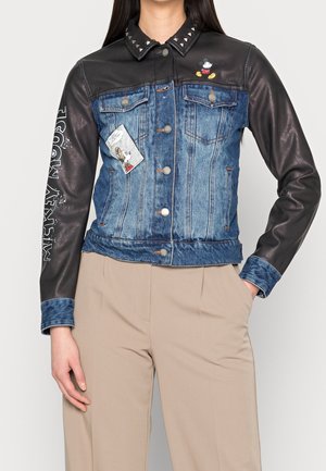 Veste en cuir noir et denim avec col clouté, patchs Mickey Mouse sur le devant et la manche, portée avec un pantalon beige.