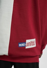 Tissu bloqué rouge et blanc avec le logo "eckō unltd." en bleu, blanc et rouge près de l'ourlet inférieur.