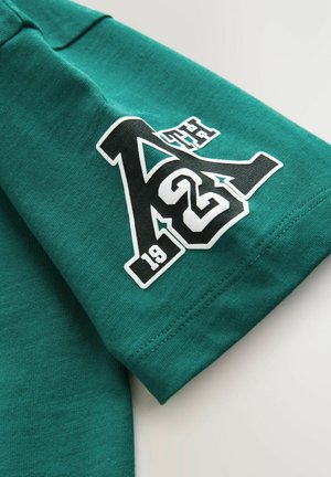 T-shirt en coton vert à manches courtes. Il présente un grand graphisme blanc et noir avec l'inscription "A2 19" sur la manche. Texture douce, design décontracté.