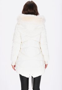 Geacă matlasată albă, cu glugă din blană sintetică, model matlasat și talie ajustabilă cu șnur. Mâneci lungi și tiv posterior ușor mai lung.