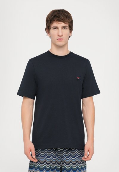T-shirt en coton bleu marine à manches courtes. Présente un petit logo de vague rouge sur la poitrine. Porté avec un short à motifs en design zigzag.