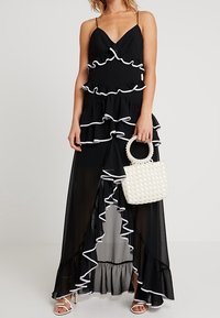 Schwarzes Kleid mit gerüschten Stufen und weißen Akzenten, mit Spaghetti-Trägern und asymmetrischem Saum, kombiniert mit einer weißen Perlenhandtasche.