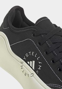 Baskets basses noires avec semelle blanche épaisse, coutures blanches et logo blanc circulaire avec texte sur le panneau latéral.