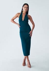 Teal halterneck-mekko, jossa on syvä V-pääntie, vyötäisille istuva siluetti ja polvipituinen helma. Valmistettu sileästä kankaasta, jossa on poimutettu yksityiskohtia.