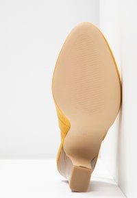 Żółty zamszowy but na wysokim obcasie z teksturowaną podeszwą, spiczastym czubkiem i cienkim obcasem, umieszczony na białym tle.