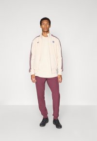 Beżowa kurtka dresowa z czerwonymi paskami, logo na piersi, w połączeniu z beżową koszulą i bordo spodniami. Czarne trampki dopełniają strój.