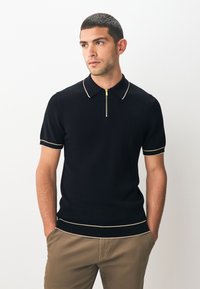 Zwart geribd poloshirt met korte mouwen, ritssluiting bij de hals en contrasterende gele biezen bij de kraag en manchetten. Draagt bij een kaki broek.