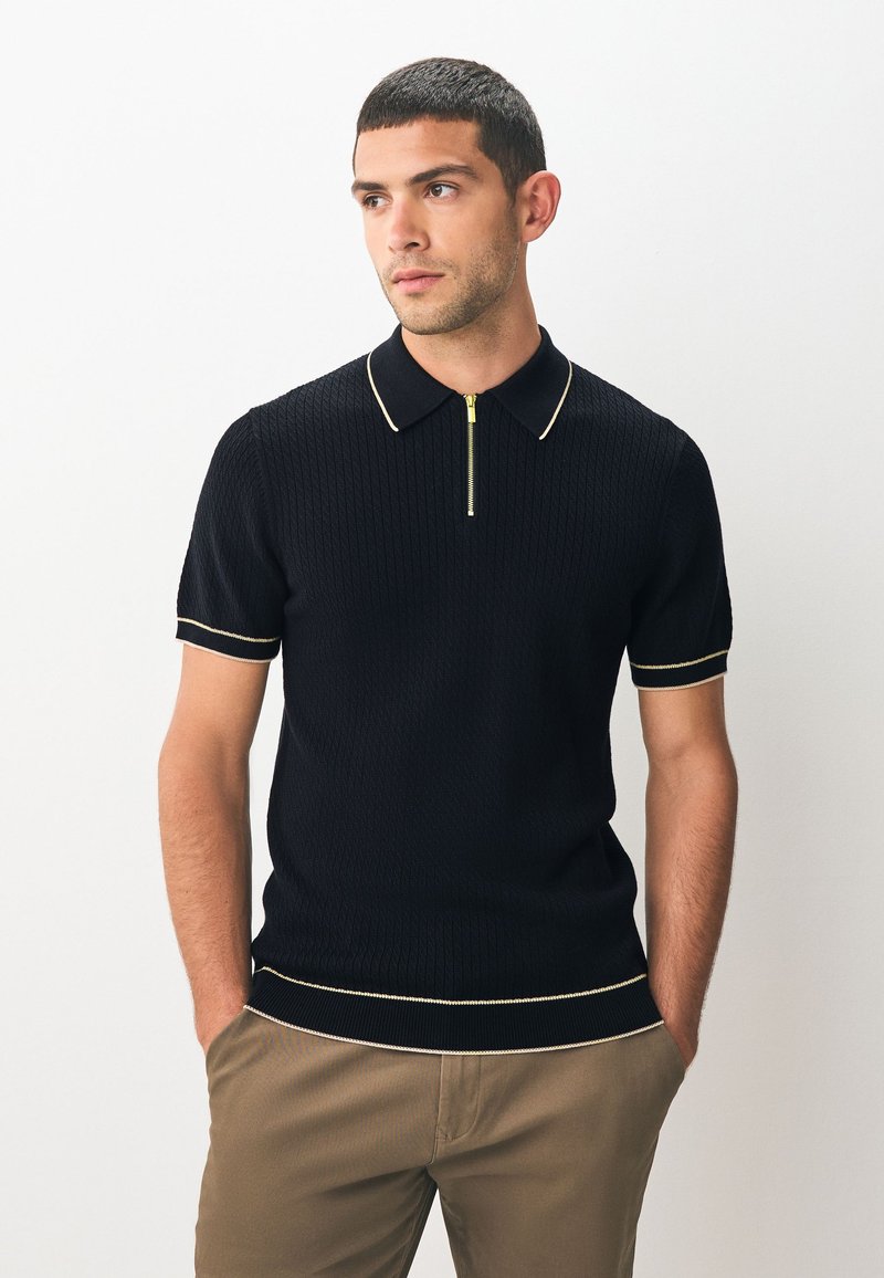 Zwart geribd poloshirt met korte mouwen, ritssluiting bij de hals en contrasterende gele biezen bij de kraag en manchetten. Draagt bij een kaki broek.