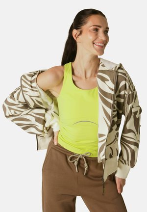 Femeie zâmbitoare cu păr lung și închis la culoare poartă un maiou neon galben, pantaloni maro cu șnur și o jachetă bej cu imprimeu de zebră așezată pe umeri.