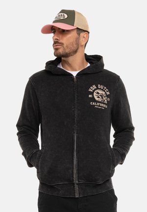 Homme portant un sweat à capuche noir zippé avec le logo "Von Dutch California" et une casquette trucker beige et rose, regardant vers la gauche.