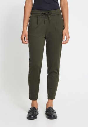Femme portant un pantalon fuselé vert olive avec des poches latérales, des mocassins noirs et un haut noir sur un fond uni.