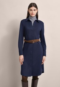 Marineblaues Kurzarmkleid mit langen Ärmeln, einem Kragen und einem taillierten Gürtel. Getragen über einem schwarz-weißen gestreiften Rollkragenpullover. Glatte Stoffoberfläche.