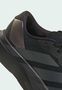 Zapatilla deportiva negra con parte superior de tela texturizada, acento moldeado en el talón y una suela elegante y ligera con una inscripción de marca visible.