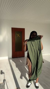 Eine Person hält ein Paar grüne Hosen in einem sonnenbeschienenen leeren Raum mit weißen Wänden, einer Holztür und einem hellen Holzboden hoch.
