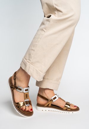 Sandales en bronze avec des accents blancs, dotées d'une semelle plate, d'un orteil ouvert et de deux détails décoratifs de boucles. Portées avec un pantalon côtelé beige.