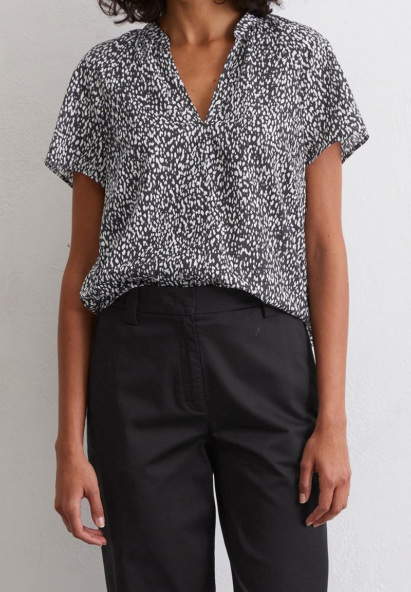 Femme portant un chemisier à manches courtes noir et blanc à pois, rentré dans un pantalon noir taille haute, debout contre un mur neutre.