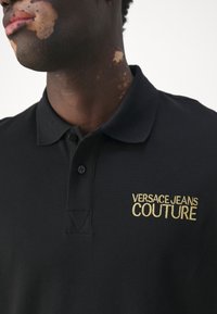 Polo noir en tissu texturé, avec un logo doré brodé "VERSACE JEANS COUTURE" sur le côté gauche. Fermeture à deux boutons.