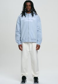 Felpa zip blu chiaro con tasche frontali e logo ricamato bianco, abbinata a pantaloni jogger bianchi e sneaker nere con dettagli bianchi.