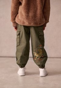 Olivgröna cargobyxor i bomull med en broderad tiger och blommönster. Tighta muddar och sidfickor. Kombineras med en beige fleecejacka.