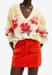 Pull en maille avec col en V, présentant un motif floral en rouge, jaune et vert. Associé à une mini-jupe en denim orange vif avec des poches.