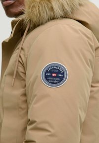 Giacca imbottita beige con cappuccio in pelliccia sintetica. Presenta una toppa rotonda blu con scritte bianche che recitano "JACK & JONES EST. 1990" sulla manica.