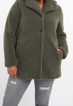 Personne portant un manteau polaire texturé vert avec des boutons et un jean gris déchiré, main dans la poche du manteau, debout devant un fond blanc.