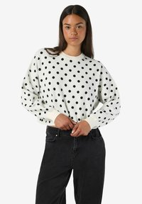 Balts džemperis ar melniem punktiņiem, apaļa kakla izgriezums, nolaižamas plecu šuves un ribota aproce. Saskaņots ar augsto vidukli melnām džinsām.