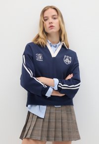 Felpa navy con strisce bianche ed emblemi, indossata sopra una camicia a righe azzurro chiaro; abbinata a una gonna plissettata a scacchi in toni di marrone.