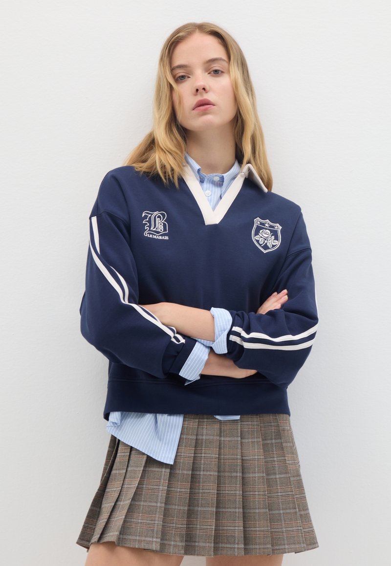 Felpa navy con strisce bianche ed emblemi, indossata sopra una camicia a righe azzurro chiaro; abbinata a una gonna plissettata a scacchi in toni di marrone.