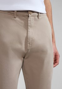 Chinos beige realizzati in tessuto leggero con una texture morbida. Presentano una chiusura frontale con bottone e tasche laterali.