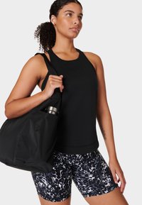 Vrouw in een zwart mouwloos shirt en een patroon op sportshorts, houdt een zwarte sporttas vast met een roestvrijstalen waterfles erin.