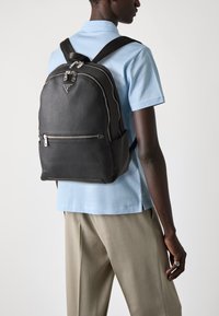 Sac à dos en cuir texturé noir avec des zips argentés et un logo triangulaire. Présente des bretelles rembourrées pour le confort et une poche zippée à l'avant.