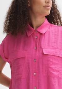 Maglietta rosa a maniche corte con finitura testurizzata, caratterizzata da un colletto, due tasche frontali e bottoni argentati lungo la parte anteriore.