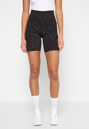 Mujer con pantalones deportivos negros de tiro alto con un sutil estampado de cebra, calcetines blancos hasta el tobillo, zapatillas blancas y una camiseta blanca sin mangas.