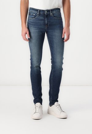 Mann trägt dunkelblaue Skinny-Jeans und weiße Turnschuhe, steht vor einem schlichten weißen Hintergrund.