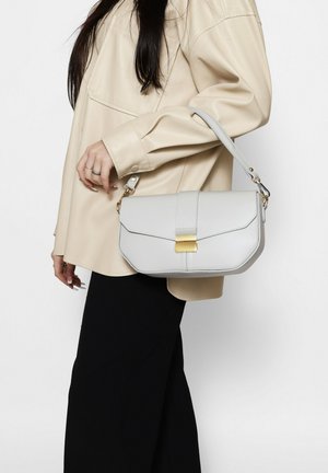 Femme en veste oversize beige et jupe noire tenant un sac à main gris clair avec fermeture dorée sur fond blanc.
