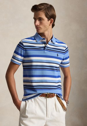 Homme portant un polo rayé bleu et blanc rentré dans un pantalon blanc avec une ceinture tressée marron, regardant sur le côté.