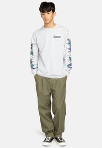 Junger Mann steht mit den Händen in den Taschen, trägt ein hellgraues Langarmshirt mit bunten Ärmelgrafiken, olivfarbene Hosen und schwarze Sneakers.