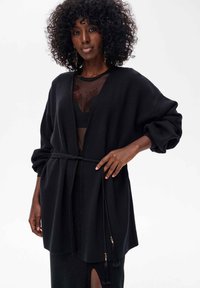 Czarny kardigan o luźnym kroju, z długimi rękawami i plecionym paskiem. Charakterystyczne detale to akcenty z chwostami oraz teksturowana tkanina.