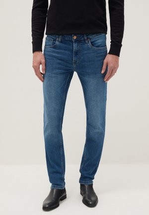 Man draagt een slim-fit blauwe spijkerbroek, een zwart shirt met lange mouwen en zwarte leren schoenen, staand tegen een eenvoudige lichte achtergrond.