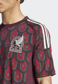Camisa marrón con tejido estampado en verde y rojo, que presenta un emblema de México y tres rayas blancas en el hombro.