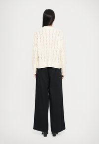 Pull crème tricoté épais avec un motif en V, associé à un pantalon noir ample, vu de l'arrière. Des chaussures à talons noires sont visibles.