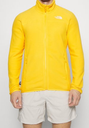 Mand iført en klar gul fleecejakke med fuld lynlås og lyse cargo-shorts, stående mod en ensfarvet baggrund.