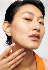 Närbild av en person med kort svart hår, lätt solbrun hy och naturlig sminkning. De rör vid sin nacke med en hand som har nude naglar.