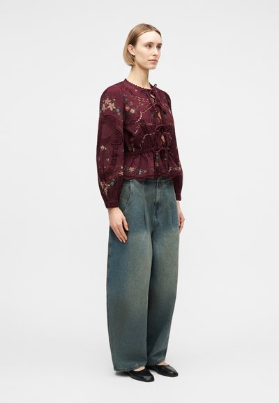 Γυναίκα που φοράει μπορντό μπλούζα με λουλουδάτο κέντημα και κορδόνια, με μακριά μανίκια και φαρδιά ξεβαμμένα μπλε τζιν παντελόνια, στέκεται μπροστά σε λευκό φόντο.
